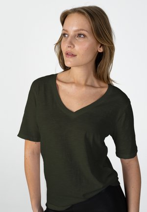 Woman with light brown hair wearing a dark green V-neck short sleeve shirt, looking slightly to her left against a plain white background.
