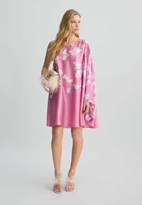 Abito in satin rosa con design asimmetrico e motivi floreali bianchi. Abbinato a una borsa a tracolla con piume e sandali nude con tacco.