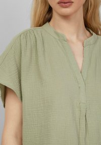 Camicia in tessuto verde chiaro con texture, colletto a V, maniche corte e sottili dettagli arricciati vicino alle spalle.