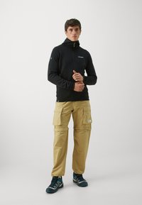 Crna fleece jakna na zatvarač s kapuljačom, s zelenim detaljima. Bež cargo hlače s višestrukim džepovima. Tamni cipelice upotpunjuju outfit.
