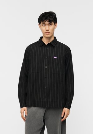 Jonge man draagt een zwart gestreept overhemd met knopen en borstzakken en een grijze broek, tegen een effen lichte achtergrond.