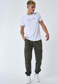 Giovane uomo con capelli biondi corti che indossa scarpe da ginnastica bianche, pantaloni cargo verde oliva e una maglietta bianca con la scritta "Project x Paris", in piedi e con lo sguardo rivolto verso il basso.