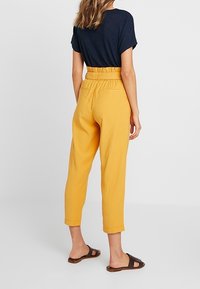 Gula högmidjade byxor med bälte i midjan, avsmalnande ben och en slät textur, kombinerat med en ribbad marinblå topp och bruna sandaler.
