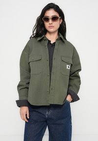 Πουκάμισο-τζάκετ σε χακί πράσινο χρώμα με δύο τσέπες στο στήθος, κλείσιμο με κουμπιά και άνετη εφαρμογή, φορεμένο πάνω από μαύρο ριμπ πλεκτό πουκάμισο.