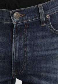 Vaqueros de mezclilla azul oscuro con cierre de botón, costuras en tono cobre y una etiqueta metálica de marca, con un corte recto y diseño clásico.