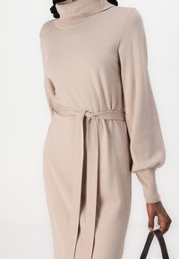 Vestito beige a maglia con collo alto, maniche lunghe a sbuffo e vita con cintura. Il tessuto appare morbido e setoso, ideale per essere indossato a strati.