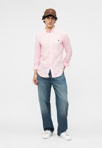 Jauns vīrietis valkā gaiši rozā pogājamu kreklu, brūnu cepuri, zilu plašu džinsu bikses un baltas sporta kurpes, stāvot pret vienkrāsainu baltu fonu.