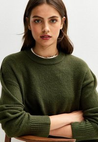 Pull vert olive en tissu doux avec un col et des poignets côtelés, présenté sur un modèle les bras croisés contre un arrière-plan clair.