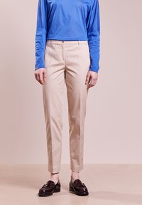 Osoba w beżowych spodniacjach o kroju dopasowanym, niebieskiej bluzce z długim rękawem i ciemnobrązowych mokasynach, stojąca na tle jasnoróżowym.