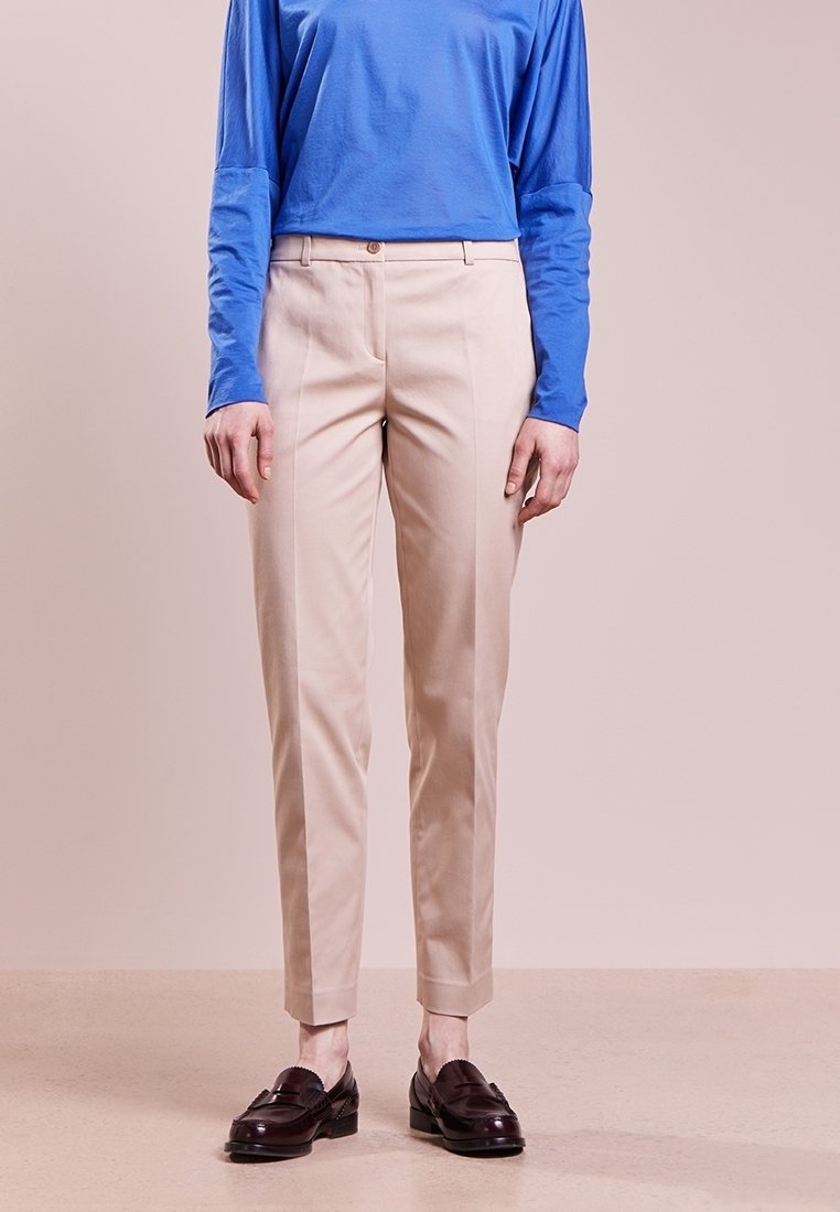 Osoba w beżowych spodniacjach o kroju dopasowanym, niebieskiej bluzce z długim rękawem i ciemnobrązowych mokasynach, stojąca na tle jasnoróżowym.