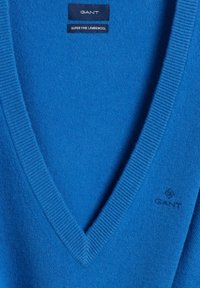 Niebieski sweter z owczej wełny, z dekoltem w serek. Posiada prążkowany kołnierz i mankiety. Logo marki jest wyhaftowane na dolnej części przodu. Miękka faktura.