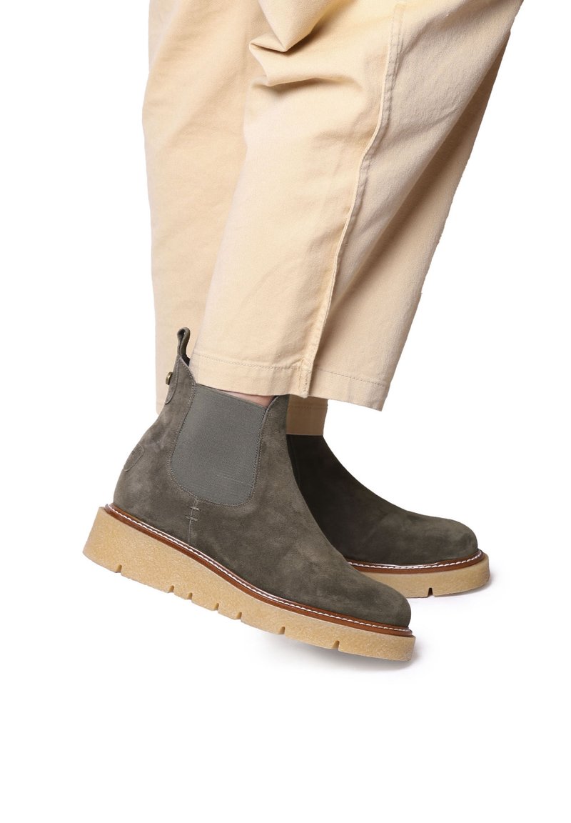 Bottes basses en daim vert olive avec panneaux latéraux élastiques, bout rond et semelle en caoutchouc texturée. Coutures détaillées visibles.