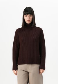 Frau mit glattem, dunklem Haar, die einen dunkelbraunen, gerippten Rollkragenpullover und helle beige Hose trägt, vor einem weißen Hintergrund stehend.