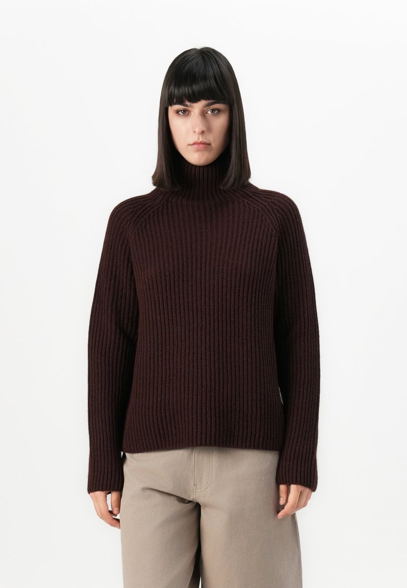 Frau mit glattem, dunklem Haar, die einen dunkelbraunen, gerippten Rollkragenpullover und helle beige Hose trägt, vor einem weißen Hintergrund stehend.