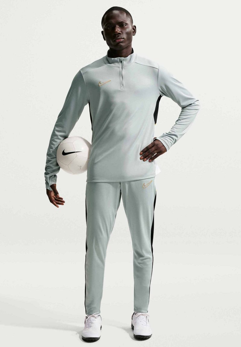 Άνδρας με γκρι φόρμα Nike κρατά με το ένα χέρι μια λευκή μπάλα ποδοσφαίρου Nike, στέκεται μπροστά σε απλό φόντο.