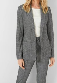 Femme portant un blazer gris à carreaux et un pantalon taille haute assorti avec un cordon de serrage, assortis à un haut blanc.