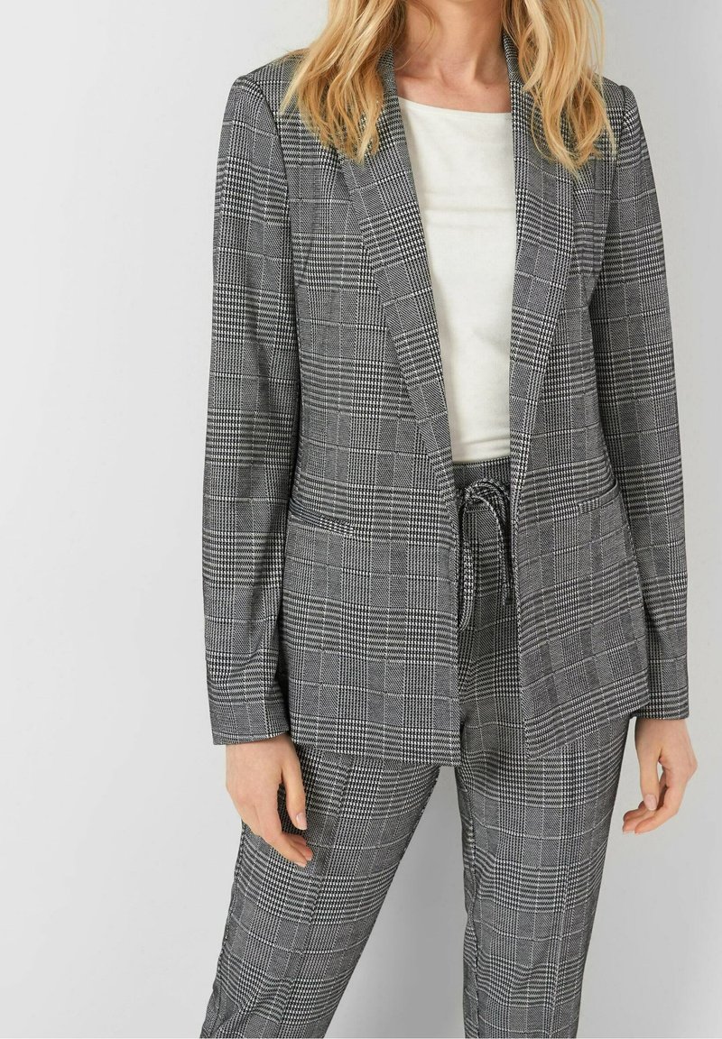Femme portant un blazer gris à carreaux et un pantalon taille haute assorti avec un cordon de serrage, assortis à un haut blanc.