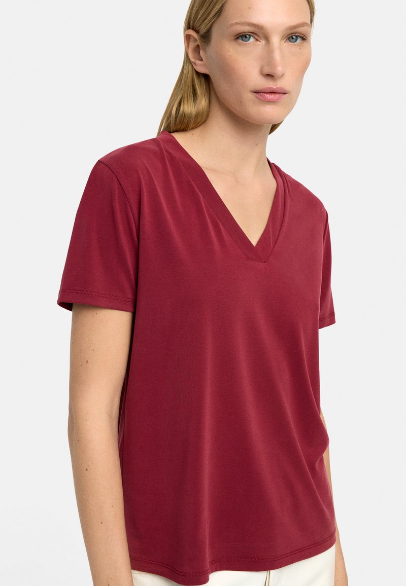 Czerwona koszulka z dekoltem w szpic wykonana z gładkiego materiału, z krótkimi rękawami i luźnym krojem, z minimalnymi szwami i prostym wzorem.