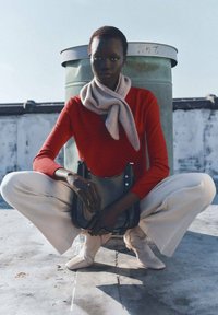 Roter Langarmpullover, hellgrauer Schal, cremefarbene weite Hose und weiche beige Ankle Boots. Modell hockt mit einer schwarzen Handtasche.