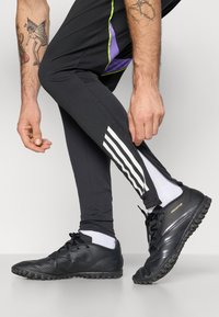 Zwarte sportieve schoenen hebben een gestructureerde bovenkant, een ronde neus en een rubberen zool. Een man draagt zwarte leggins met witte strepen en paarse accenten.