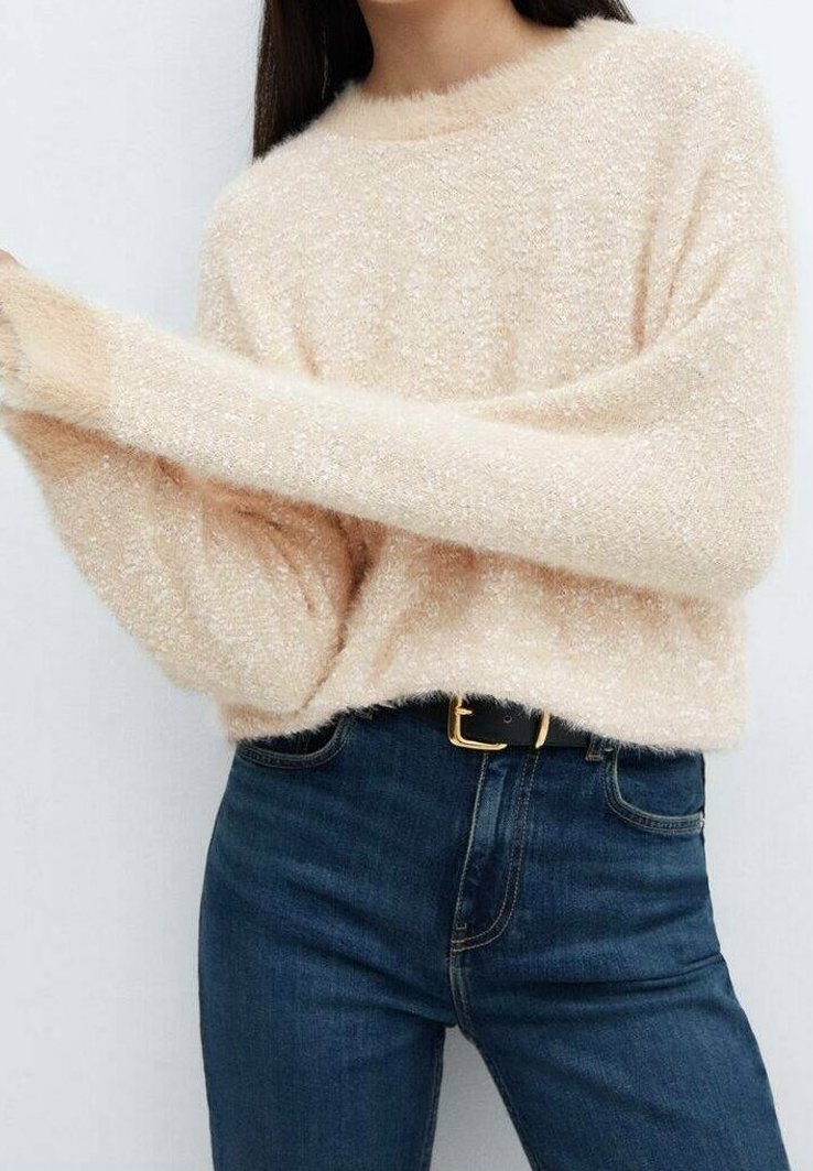 Femme portant un pull en maille crème duveteux à manches longues et un jean bleu foncé taille haute avec une ceinture noire à boucle dorée.