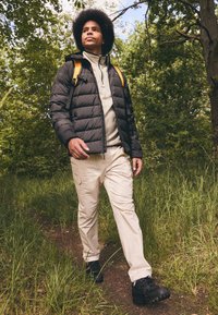 Svart puffjacka, ljusgrå fleece under, beige cargo-byxor och svarta vandringskängor. Gula remmar på ryggsäcken, grön växtlighet i bakgrunden.