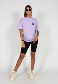 Lavender oversized t-shirt med sort grafisk logo, kombineret med sorte cykelshorts og beige sneakers, sat mod en ensfarvet hvid baggrund.