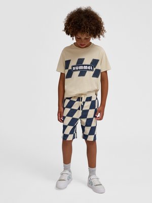 Dziecko o kręconych włosach, ubrane w beżowy t-shirt z logo "Hummel" i kraciaste spodenki, patrzy w dół, stoi na jednolitym tle.