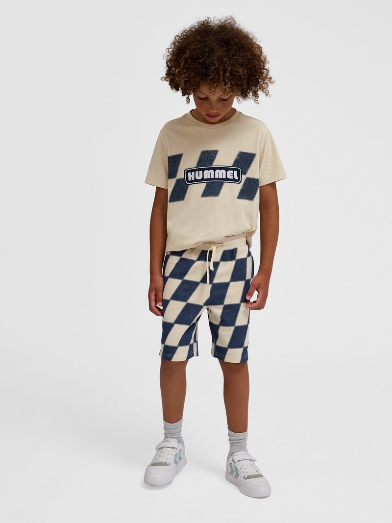 Dziecko o kręconych włosach, ubrane w beżowy t-shirt z logo "Hummel" i kraciaste spodenki, patrzy w dół, stoi na jednolitym tle.