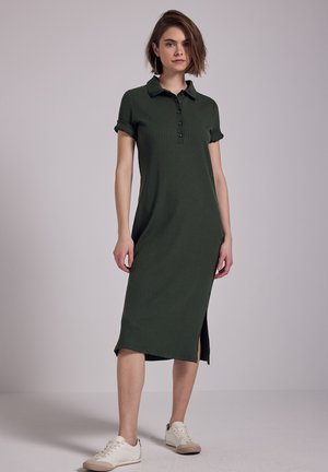Abito polo a coste verde scuro con maniche corte arrotolate, bottoni lungo la parte frontale e spacchi laterali. Abbinato a sneaker bianche.