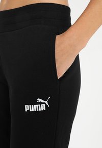 Mano en el bolsillo de pantalones deportivos negros de Puma con el logo blanco de Puma en el muslo, vista en primer plano del área de la cintura y la cadera.