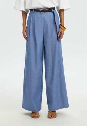 Kvinde iført højtaljede, vidde bukser i lyseblå med et brunt flettet bælte, hvid top og brune og gule armbånd.