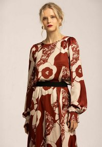 Långärmad klänning i röd och beige med blommönster och abstrakta mönster; har ett svart midjeband och rynkade ärmslut.