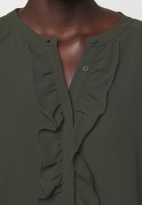 Blusa verde oscuro con un pliegue rizado en el cuello, escote redondeado y dos cierres de botón en el collar. Tejido texturizado con un acabado suave.