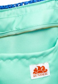 Intérieur en tissu vert menthe avec une fermeture éclair dorée et un dessus tissé bleu. Comprend une étiquette blanche avec un logo de soleil orange, marqué "SUNDEK."