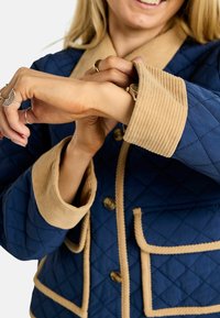 Granatowa pikowana kurtka z kontrastowym beżowym kołnierzem i mankietami z sztruksu, wyposażona w zaokrąglone kieszenie i ozdobne przeszycia. Widoczne guziki.