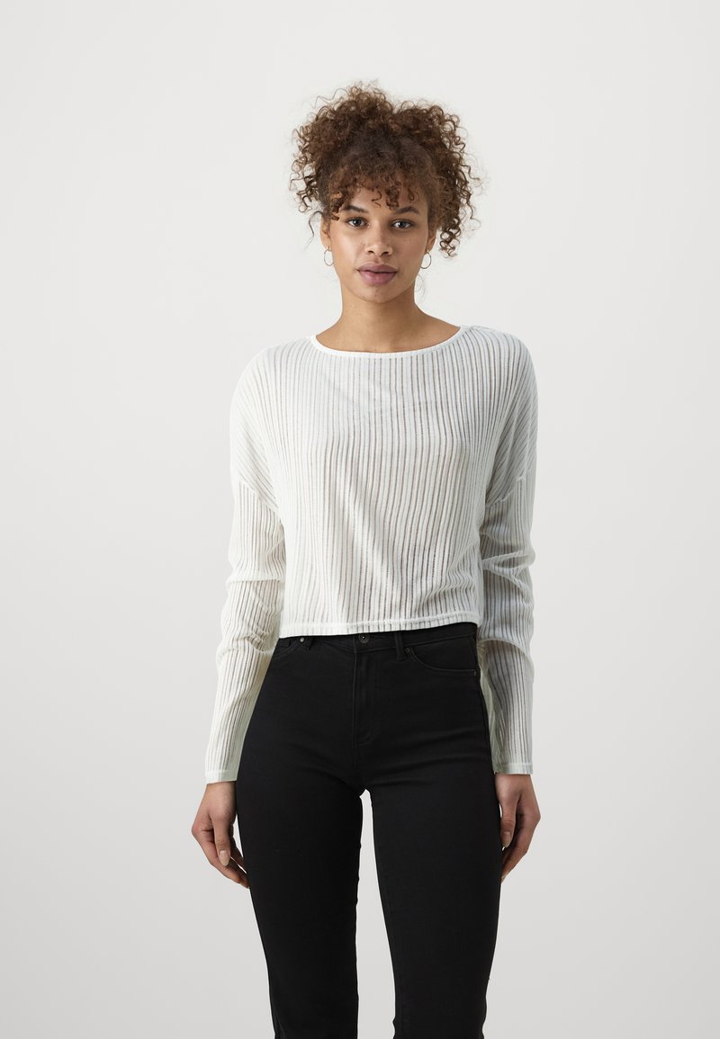 Haut à manches longues côtelé blanc avec un col bateau, associé à un jean noir taille haute. Le tissu semble léger et texturé.
