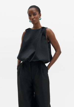 Donna che indossa un top nero in raso senza maniche con pantaloni neri a gamba larga, posando con le mani in tasca e orecchini a cerchio color turchese.