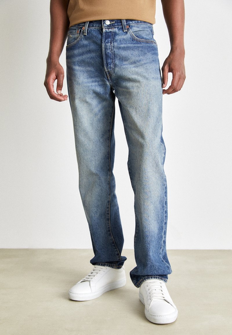 Jeans rectos de color azul claro, confeccionados en denims, con un lavado desgastado, diseño de cinco bolsillos y costuras en contraste, combinados con zapatillas blancas.