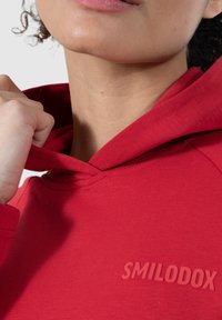 Hoodie en coton rouge avec une poche avant, capuche à cordon et un petit logo en relief "SMILODOX" sur la poitrine. Texture lisse, design décontracté.