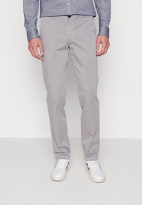 Uomo che indossa pantaloni slim-fit grigio chiaro, sneakers bianche con dettagli neri e una maglietta blu a maniche lunghe, in piedi su un pavimento grigio uniforme.