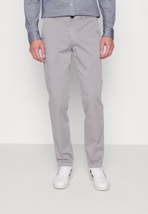 Uomo che indossa pantaloni slim-fit grigio chiaro, sneakers bianche con dettagli neri e una maglietta blu a maniche lunghe, in piedi su un pavimento grigio uniforme.