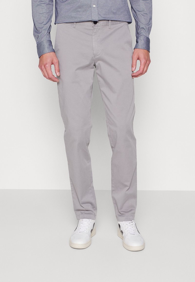 Uomo che indossa pantaloni slim-fit grigio chiaro, sneakers bianche con dettagli neri e una maglietta blu a maniche lunghe, in piedi su un pavimento grigio uniforme.