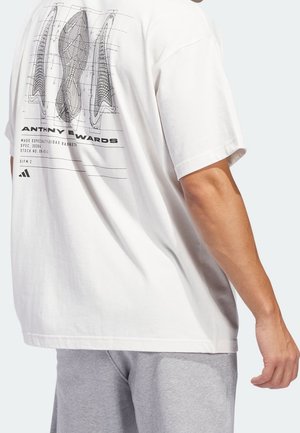 Weißes T-Shirt mit schwarzen technischen Zeichnungen von Schuhsohlen und dem Text "Anthony Edwards", getragen von einer Person in grauen Shorts, die Rücken und rechten Arm zeigt.
