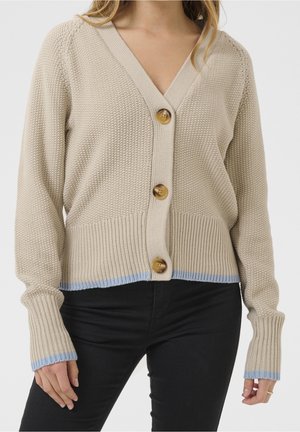 Beige strikket cardigan med tre store brune knapper og lyseblå kanter på ærmer og kant, båret med sorte bukser.