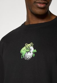 Μαύρο βαμβακερό t-shirt με γραφικό ενός πράσινου πλάσματος πάνω σε λευκή μπάλα του γκολφ, με λεπτομερείς λεπτομέρειες και στρογγυλή λαιμόκοψη.