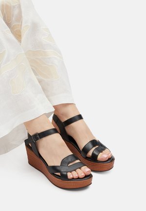 Schwarze Lederkeilsandalen mit Riemendesign, verstellbarer Knöchelschnalle und einer gerillten braunen Plateausohle. Glatte Textur.