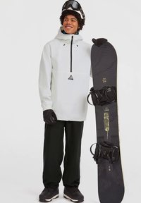 Νέος άνθρωπος φορώντας λευκή ζακέτα, μαύρα παντελόνια χιονιού, γάντια και κράνος κρατάει μαύρη σανίδα snowboard ενώ στέκεται σε λευκό φόντο.