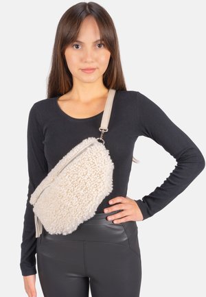 Sac bandoulière en fausse peau de mouton couleur crème avec une fermeture éclair argentée, une sangle réglable et une surface texturée. Forme arrondie avec un détail à franges sur le côté.