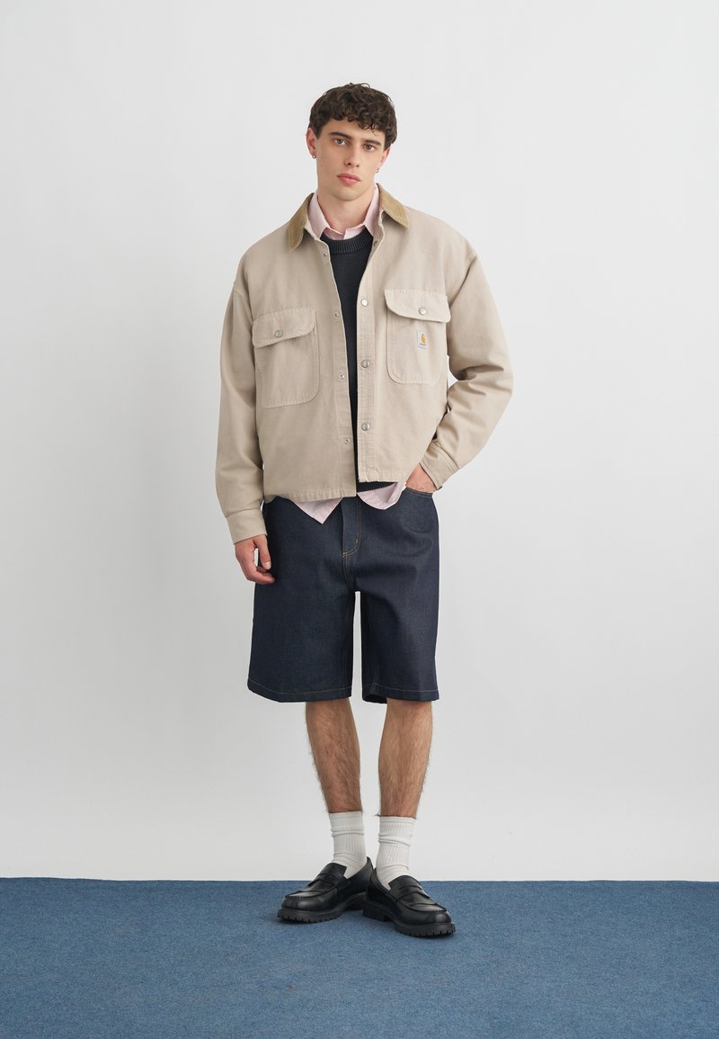 Joven con una chaqueta beige sobre un suéter negro y camisa rosa, pantalones cortos azul marino, calcetines blancos y mocasines negros, de pie sobre una alfombra azul.